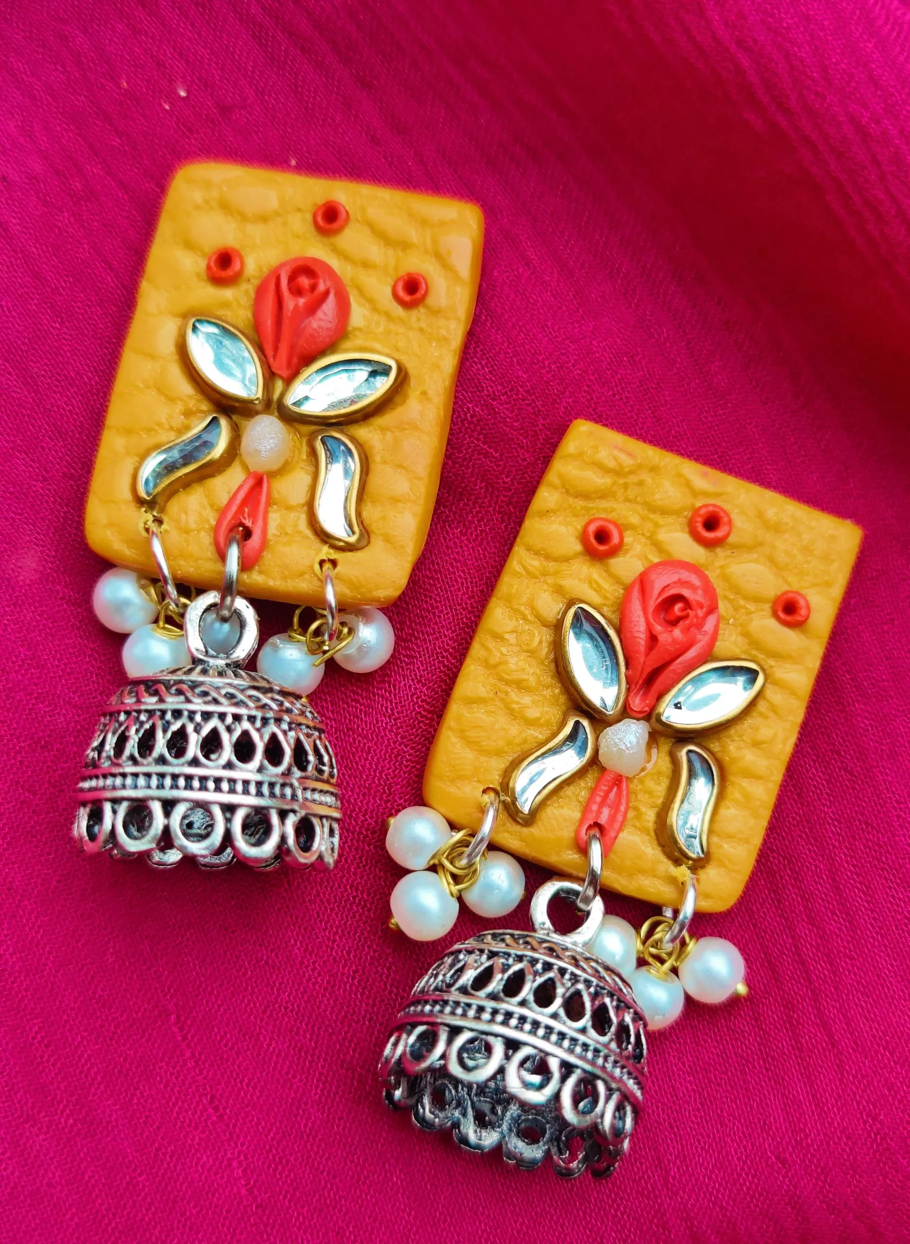 These vibrant yellow jhumka earrings are a fun mix of tradition and modern style. The square-shaped top has a textured surface, decorated with a colorful rosette motif. The earrings are finished with silver oxidized jhumkas and tiny pearl drops, adding a delicate touch.
