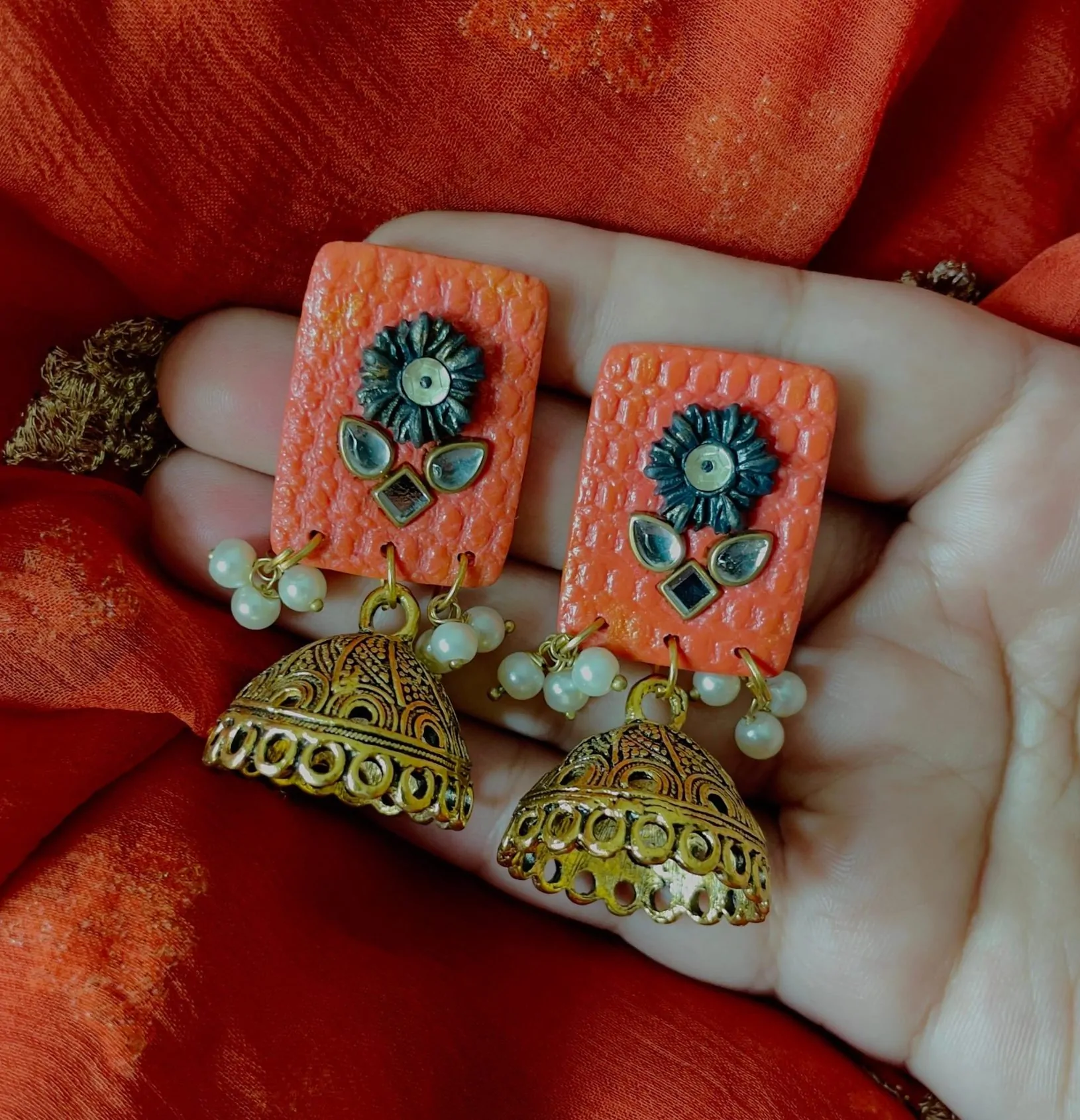 These rust-orange jhumka earrings are a beautiful mix of traditional and contemporary design. The earrings feature a rectangular textured resin top with a rich rust-orange finish, giving them a unique handcrafted look. The top is decorated with black floral metal embellishments and tiny mirror accents, adding a touch of vintage charm.