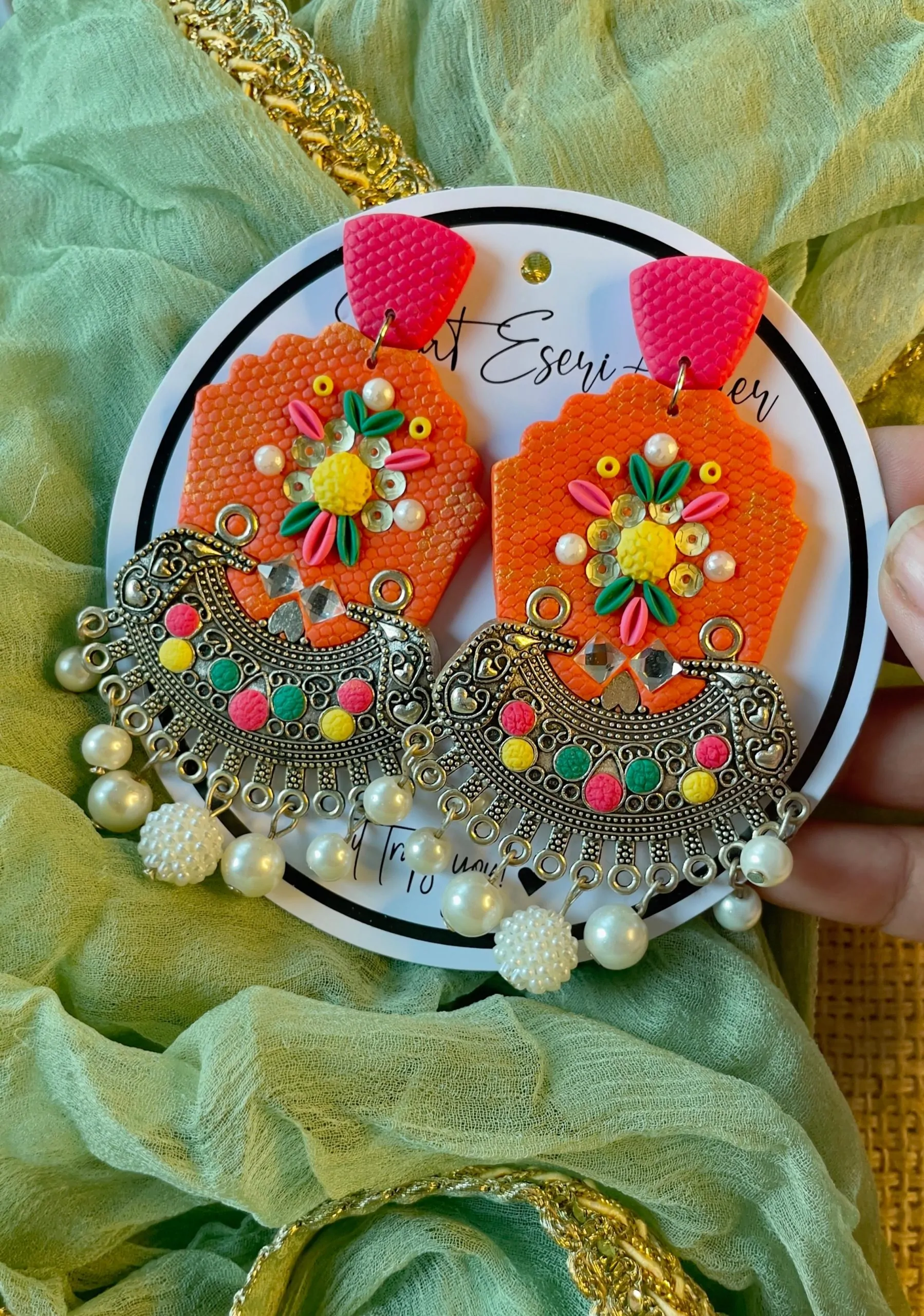A vibrant and intricate blend of traditional and contemporary design elements, our antique silver crescent-shaped earring features a textured orange base with a floral embellishment made of colorful beads and sequins in pink, yellow, green, and white.