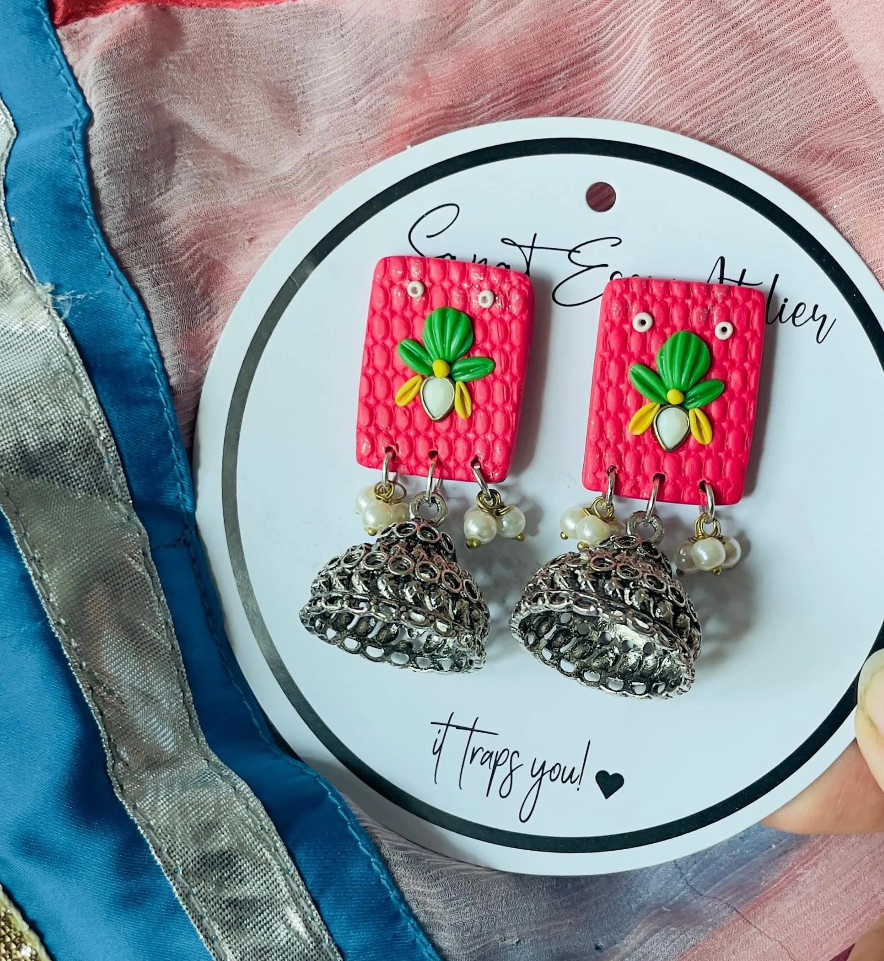 These vibrant pink jhumka earrings are a fun mix of tradition and modern style. The square-shaped top has a textured surface, decorated with a colorful floral motif in green, yellow, and white. The earrings are finished with silver oxidized jhumkas and tiny pearl drops, adding a delicate touch.