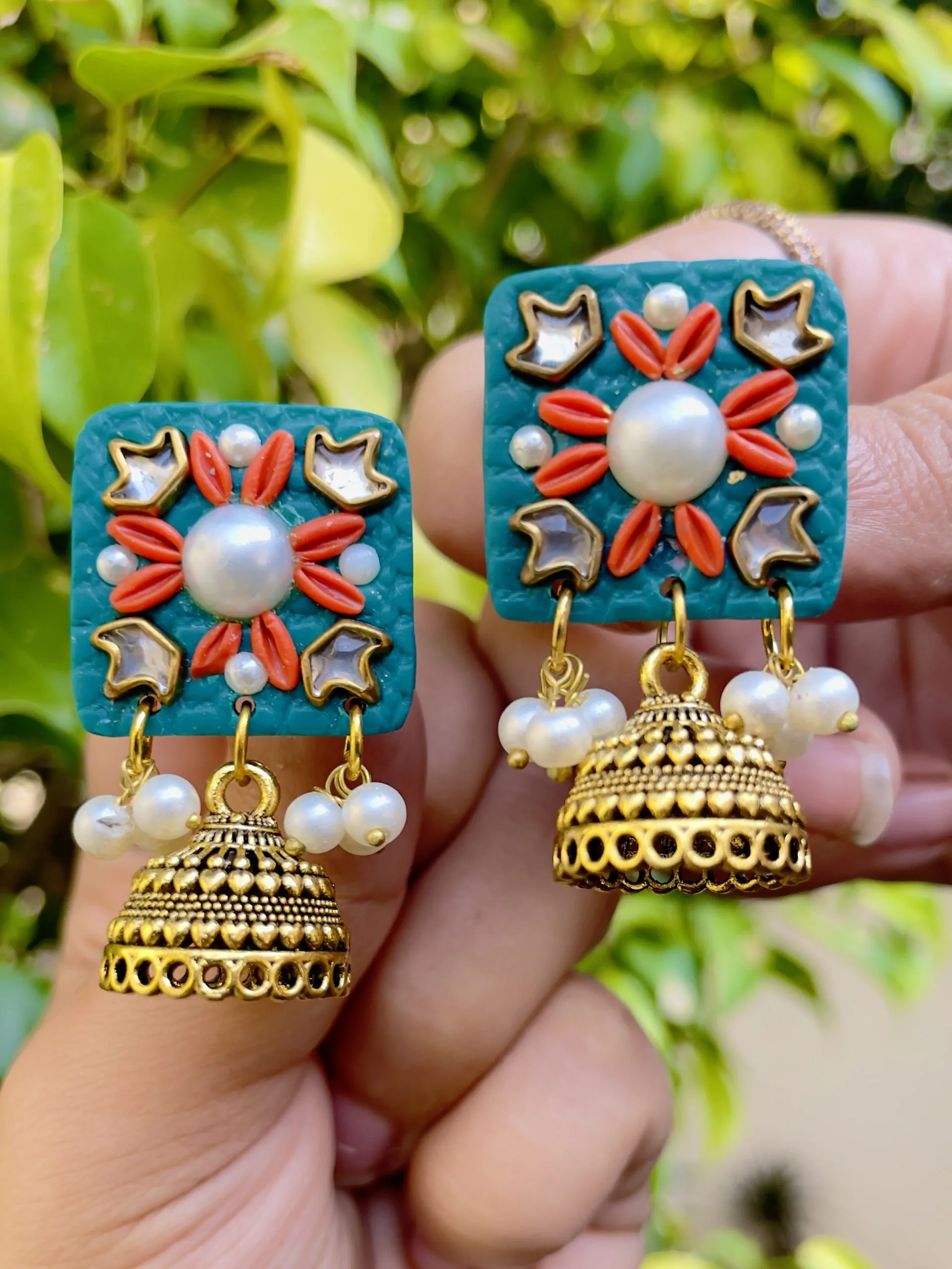 Teal square textured earrings with coral floral accents, gold Jhumka bells, and white pearls.