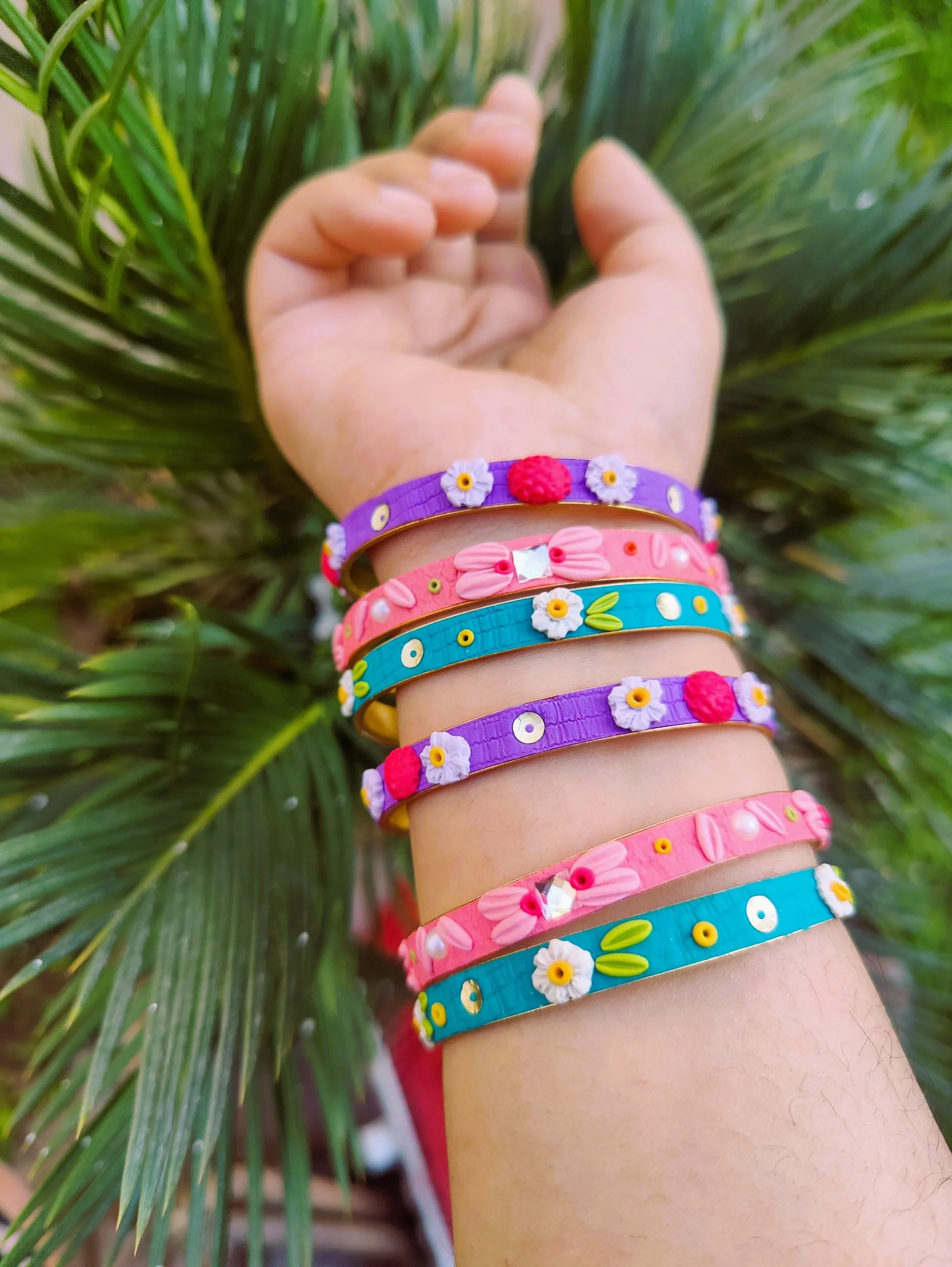 A stack of colorful artisan bangles featuring 3D clay flowers, gold thread work, and shimmering sequin accents.