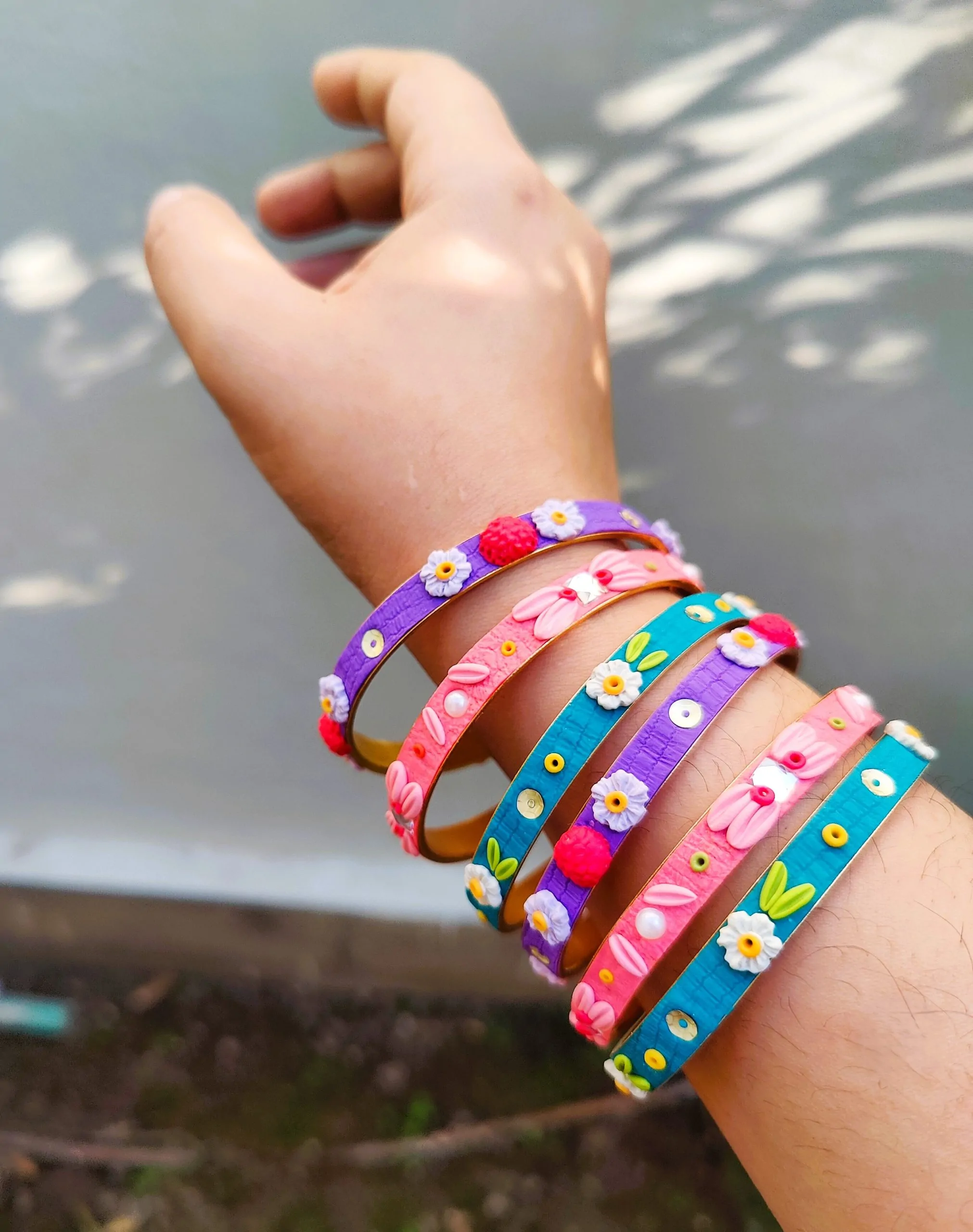 A stack of colorful artisan bangles featuring 3D clay flowers, gold thread work, and shimmering sequin accents.