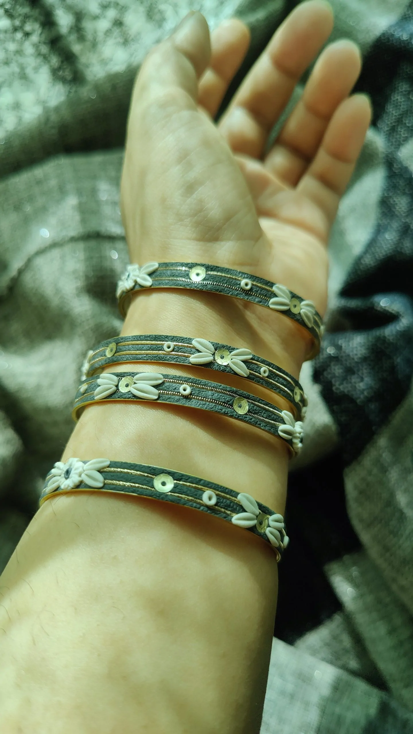IMG20240502153528 Artisan stack of charcoal grey textured bangles featuring hand-sculpted white clay flowers, silver sequins, and gold metallic threading."