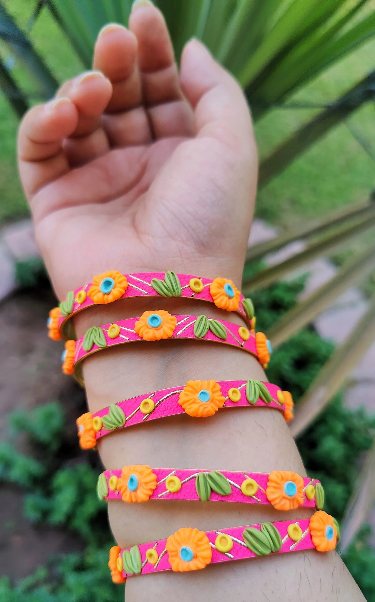 IMG_20240419_131356 Artisan stack of magenta pink textured bangles featuring hand-sculpted orange clay flowers, green leaves, and gold metallic embroidery.