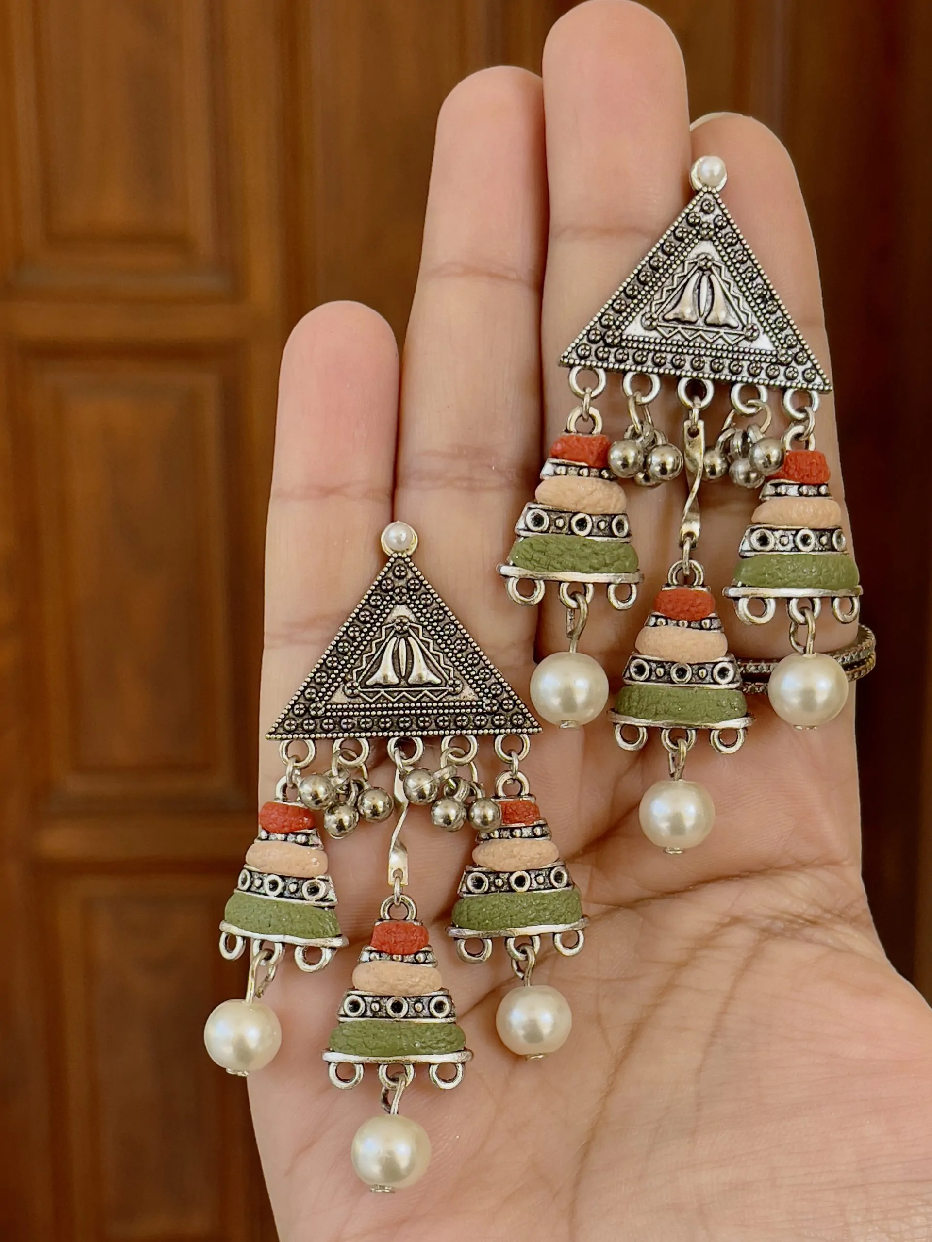 Handcrafted oxidized silver triangular earrings featuring a triple-tier tassel of terracotta red, peach, and olive green textured beads with pearl drops.